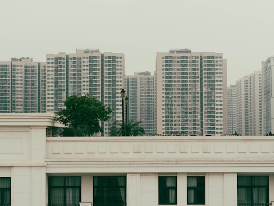 A series of high-rise apartment buildings dominates the skyline, with uniform design and numerous windows. In the foreground, there is a white building with a flat roof and a contrasting green tree. This urban scene suggests dense population and modern city living.