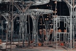A construction site featuring large, intricate metal structures resembling trees. The area is lit with artificial lighting, casting shadows and highlighting the complex framework. Surrounding the structures are various construction materials and equipment.