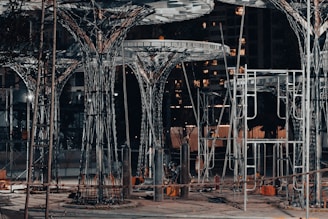 A construction site featuring large, intricate metal structures resembling trees. The area is lit with artificial lighting, casting shadows and highlighting the complex framework. Surrounding the structures are various construction materials and equipment.