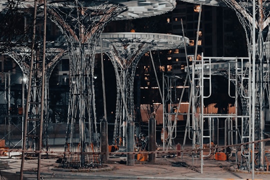 A construction site featuring large, intricate metal structures resembling trees. The area is lit with artificial lighting, casting shadows and highlighting the complex framework. Surrounding the structures are various construction materials and equipment.