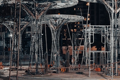 A construction site featuring large, intricate metal structures resembling trees. The area is lit with artificial lighting, casting shadows and highlighting the complex framework. Surrounding the structures are various construction materials and equipment.