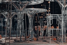 A construction site featuring large, intricate metal structures resembling trees. The area is lit with artificial lighting, casting shadows and highlighting the complex framework. Surrounding the structures are various construction materials and equipment.