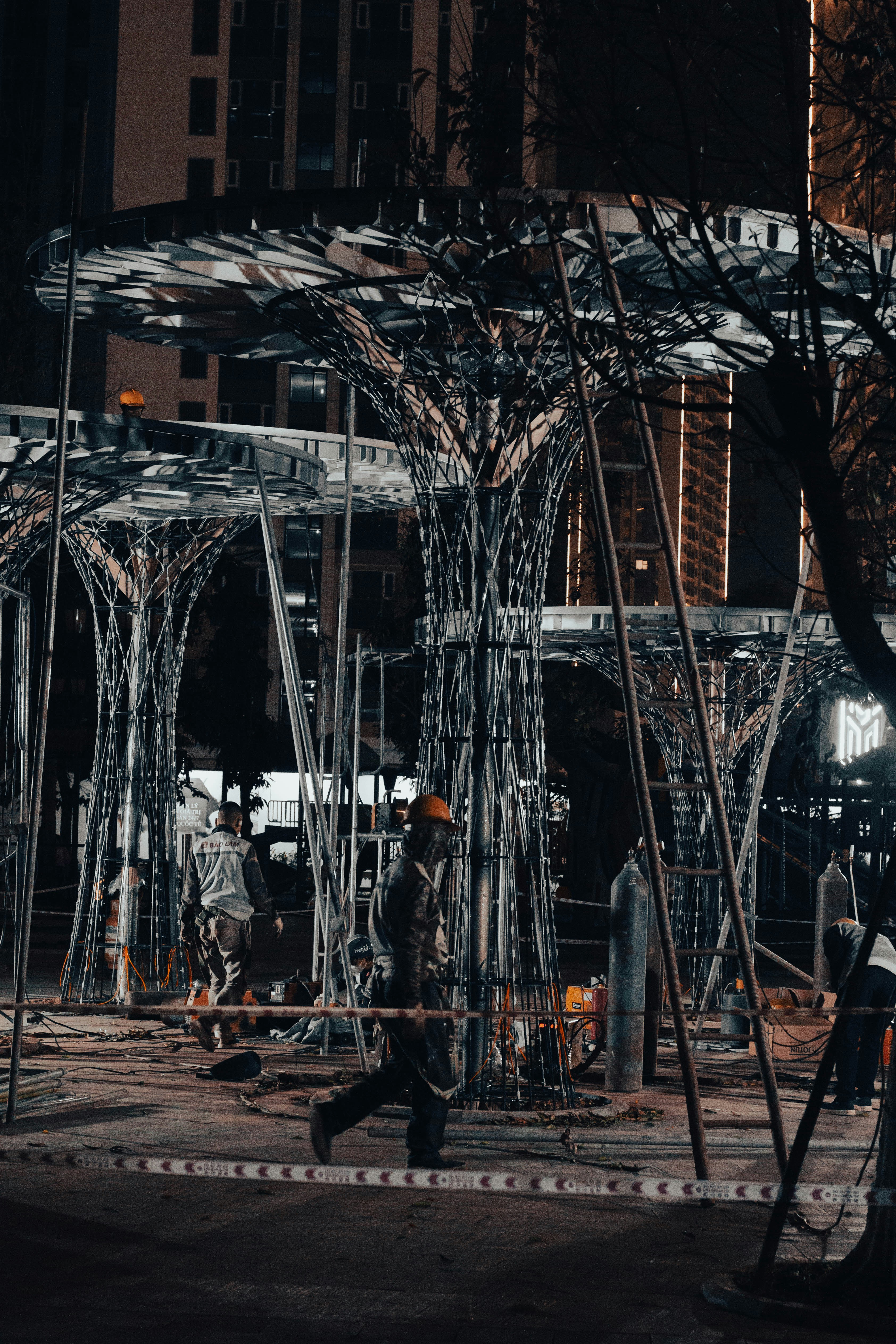 Construcción de planta industrial con estructura metálica, trabajadores con cascos, cielo oscuro con iluminación de obra