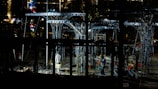 Workers are operating at a construction site during the night, surrounded by metal structures and illuminated by artificial lights. The scene is filled with scaffolding and tools, creating an industrial atmosphere.