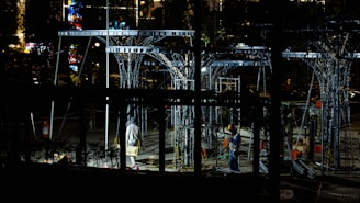 Workers are operating at a construction site during the night, surrounded by metal structures and illuminated by artificial lights. The scene is filled with scaffolding and tools, creating an industrial atmosphere.