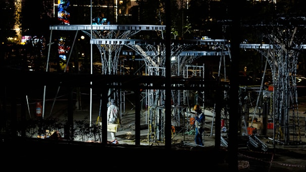 Workers are operating at a construction site during the night, surrounded by metal structures and illuminated by artificial lights. The scene is filled with scaffolding and tools, creating an industrial atmosphere.