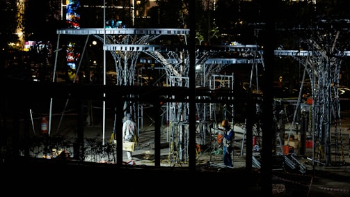 Workers are operating at a construction site during the night, surrounded by metal structures and illuminated by artificial lights. The scene is filled with scaffolding and tools, creating an industrial atmosphere.
