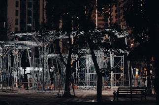 A nighttime scene of a construction site illuminated by artificial lighting. Metal scaffolding and framework structures are visible, surrounded by urban buildings. Foreground includes shadows of trees and a park bench, adding a contrasting natural element to the industrial setting.