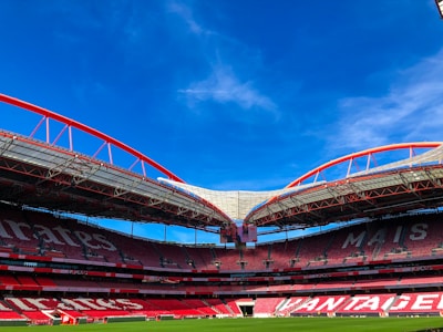 A large, empty sports stadium with distinctive red seating and a modern architectural design. The roof features red steel arches and partially transparent sections, contrasting with the bright blue sky. The grass on the field is neatly maintained.