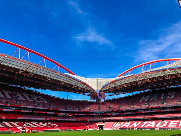 A large, empty sports stadium with distinctive red seating and a modern architectural design. The roof features red steel arches and partially transparent sections, contrasting with the bright blue sky. The grass on the field is neatly maintained.