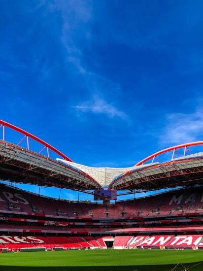 A large open football stadium with red seating and a green field under a clear blue sky. The stadium features impressive architectural structures with red and silver metallic accents.