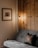 A cozy room features a grey sofa with a decorative cushion depicting a deer. Mounted on the wall is a wooden frame containing art, which portrays a rustic cabin surrounded by trees. A warm light emanates from a minimalist floor lamp beside a wooden panel wall, creating a welcoming and serene atmosphere.