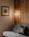 A cozy room features a grey sofa with a decorative cushion depicting a deer. Mounted on the wall is a wooden frame containing art, which portrays a rustic cabin surrounded by trees. A warm light emanates from a minimalist floor lamp beside a wooden panel wall, creating a welcoming and serene atmosphere.