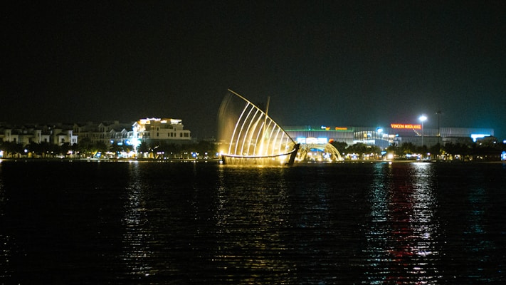 A night scene featuring a waterfront with a prominent illuminated installation that resembles a sail. The water reflects the city lights, adding a shimmering effect to the scene. In the background, several buildings are visible, including a large structure with the name 'Vincom Mega Mall' lit up.