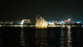 A night scene featuring a waterfront with a prominent illuminated installation that resembles a sail. The water reflects the city lights, adding a shimmering effect to the scene. In the background, several buildings are visible, including a large structure with the name 'Vincom Mega Mall' lit up.