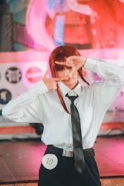 A person wearing a white shirt, black tie, and black pants, holding their hands up in a frame gesture. They stand on a stage with a pink and colorful abstract background, with a circle displaying the number 10 attached to their pants.