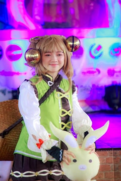 A person dressed in a colorful costume featuring a lime green and white top with black accents and puffy sleeves, accompanied by a bright green skirt. They are wearing a wig with large, ornate brown spirals and holding a plush toy that resembles a rabbit. The background is filled with neon pink and blue lights, creating a vibrant and lively atmosphere.