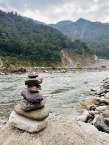 A stack of balanced stones near the edge of a flowing river with a background of forested hills and a cloudy sky. The rocks and the natural setting give a serene and harmonious feel to the image.