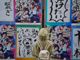 A person wearing a light green jacket and carrying a white backpack is standing in front of a wall adorned with vibrant and colorful posters. The posters feature dynamic images of people in various engaging activities, with one showing people in school uniforms holding musical instruments and another featuring bold Japanese text with graphic designs.