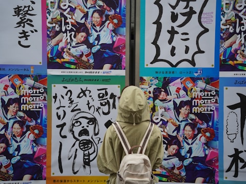 A person wearing a light green jacket and carrying a white backpack is standing in front of a wall adorned with vibrant and colorful posters. The posters feature dynamic images of people in various engaging activities, with one showing people in school uniforms holding musical instruments and another featuring bold Japanese text with graphic designs.
