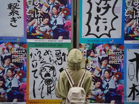 A person wearing a light green jacket and carrying a white backpack is standing in front of a wall adorned with vibrant and colorful posters. The posters feature dynamic images of people in various engaging activities, with one showing people in school uniforms holding musical instruments and another featuring bold Japanese text with graphic designs.