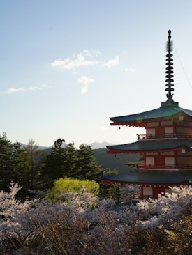 A serene landscape featuring a traditional multi-tiered pagoda partially surrounded by cherry blossom trees in full bloom. The background includes a forest and mountains under a clear sky with a few scattered clouds, suggesting a peaceful and tranquil setting.