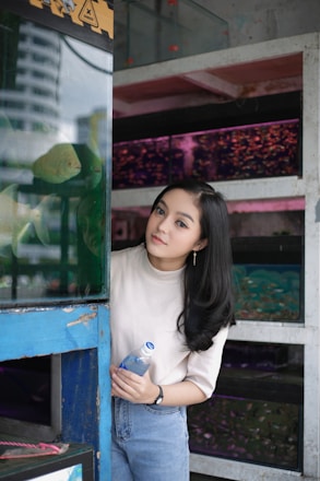 A person stands beside a blue-framed aquarium, holding a bottle of water. Several fish tanks line the background, filled with colorful fish. The setting appears to be an indoor aquarium or a similar venue.