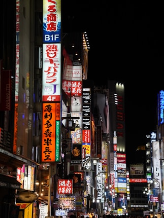 A bustling urban street illuminated by numerous colorful neon signs and billboards, featuring Japanese characters and advertising various establishments. The scene is lively and vibrant, with lights contrasting against the night sky.