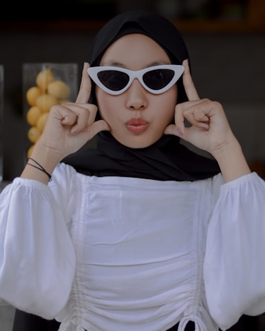 A person wearing a white blouse and black hijab is posing with their fingers forming a playful gesture near their face. They have on white cat-eye sunglasses and are pursing their lips, creating a fun and expressive mood. In the background, there is a container filled with yellow lemons.