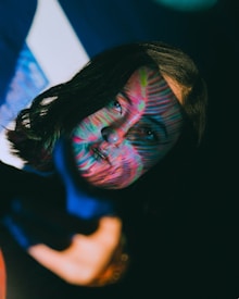 A person with dark hair is partially illuminated by colorful, patterned light projections, creating a striking visual effect across their face. The background is dark, enhancing the vividness of the colors on the person's face and surroundings.