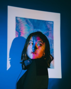 A person with dark hair is captured in a side profile, partially illuminated by vibrant, colorful light projections. These projections include shades of blue, pink, and purple, creating a striking interplay of colors on the person's face and the wall behind them. The surrounding is dark, enhancing the vividness of the colors. A pair of headphones rests around the neck.