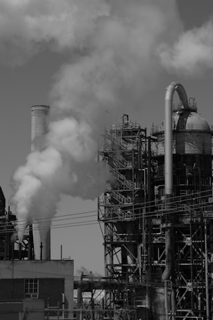 A large industrial facility with complex metal structures and tall chimneys emitting thick clouds of smoke into the sky. The setting appears to be a factory or power plant with numerous pipes and platforms. The atmosphere is industrial and mechanical.