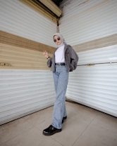 A person stands confidently in front of a textured, beige and white wall, wearing sunglasses, a gray hijab, a gray cropped jacket, a white turtleneck, light blue jeans, and black shoes. The individual poses with one hand slightly raised and the other resting at their side, exuding a casual yet stylish demeanor.