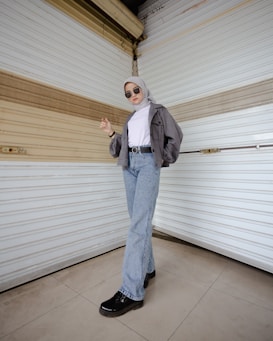 A person stands confidently in front of a textured, beige and white wall, wearing sunglasses, a gray hijab, a gray cropped jacket, a white turtleneck, light blue jeans, and black shoes. The individual poses with one hand slightly raised and the other resting at their side, exuding a casual yet stylish demeanor.