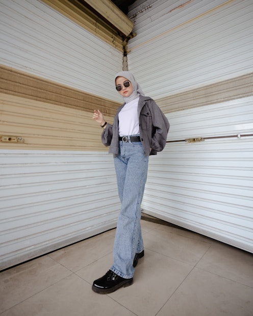 A person stands confidently in front of a textured, beige and white wall, wearing sunglasses, a gray hijab, a gray cropped jacket, a white turtleneck, light blue jeans, and black shoes. The individual poses with one hand slightly raised and the other resting at their side, exuding a casual yet stylish demeanor.