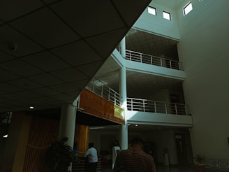 An interior view of a modern building with a multi-level, open space design. There are several floors with railings and an orange banner displaying an institution's name. The ceiling features grid-like panels. People are present, with one person in focus walking away, and another figure near a reception area surrounded by some indoor plants.