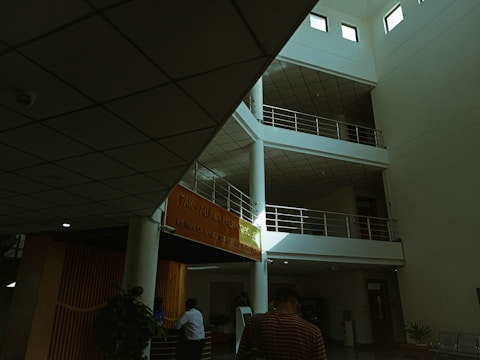 An interior view of a modern building with a multi-level, open space design. There are several floors with railings and an orange banner displaying an institution's name. The ceiling features grid-like panels. People are present, with one person in focus walking away, and another figure near a reception area surrounded by some indoor plants.
