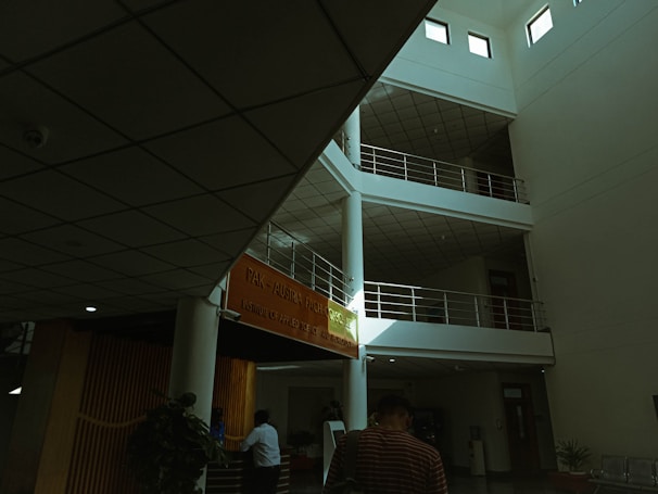 An interior view of a modern building with a multi-level, open space design. There are several floors with railings and an orange banner displaying an institution's name. The ceiling features grid-like panels. People are present, with one person in focus walking away, and another figure near a reception area surrounded by some indoor plants.