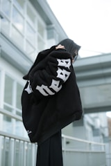 A person wearing a large black jacket with bold white lettering, standing outdoors against a modern architectural background featuring glass and metal elements. The individual appears to be adjusting their sunglasses.
