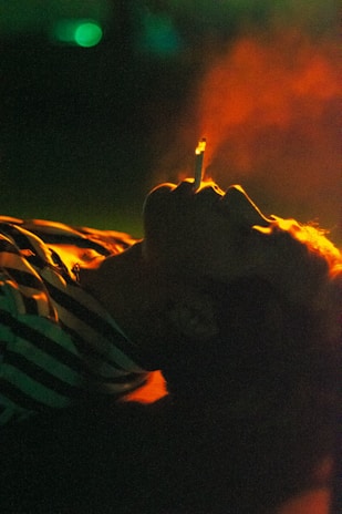 A person is lying down with a cigarette in their mouth, emitting smoke. The lighting is dim with strong hues of orange, green, and yellow, creating an intense and moody atmosphere. The person is wearing a striped garment.