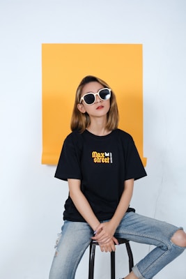 A person with shoulder-length hair is sitting on a stool in front of a yellow backdrop. They are wearing oversized black and white sunglasses, a black T-shirt with graphic text reading 'Max street,' and ripped blue jeans. The overall appearance is casual and stylish.