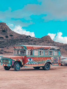 A vibrantly painted vintage bus with intricate psychedelic designs, including eyes and abstract patterns. The bus is parked on a dirt area with arid, rocky hills in the background under a bright blue sky with scattered clouds.