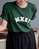 Person wearing a dark green T-shirt with the letters MXST printed on it in white. The T-shirt is paired with a black skirt and the person is leaning against a wooden railing.