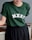Person wearing a dark green T-shirt with the letters MXST printed on it in white. The T-shirt is paired with a black skirt and the person is leaning against a wooden railing.