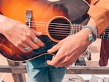 A person is holding an acoustic guitar. The person's hands are adorned with turquoise jewelry, including a ring and bracelet. The guitar is dark wood with a sunburst finish. The person is wearing a tan jacket and blue jeans.