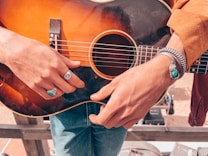 A person is holding an acoustic guitar. The person's hands are adorned with turquoise jewelry, including a ring and bracelet. The guitar is dark wood with a sunburst finish. The person is wearing a tan jacket and blue jeans.