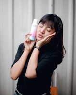 A person is gently holding a bottle of skincare product beside their face, giving an expression of contentment and calm. The background is a muted, neutral color, and the person is wearing a black top and a headband.