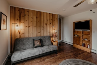 A cozy living room with a rustic vibe, featuring a wooden accent wall and a dark gray couch. A small, round table with a lamp and a decorative pillow with a deer design enhances the warm ambiance. A simple entertainment center with a television, books, and decorative items is placed against the white wall. A floor lamp with a minimalist design is positioned next to the couch, casting soft light.