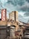 A rundown urban street scene features old buildings with various signage, including one for 'Jasco' and another for 'Jilani Steel.' Overhead, tangled utility wires crisscross the sky, which is overcast with dark clouds. In the background, a modern high-rise building contrasts with the more decrepit structures in the foreground. A parked car is visible on the right-hand side of the image.
