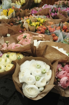 A variety of flower bouquets are arranged on display. The bouquets are wrapped in brown paper and contain different types of flowers, including roses and tulips. The colors range from white and pink to yellow and orange, creating a vibrant and lively scene.